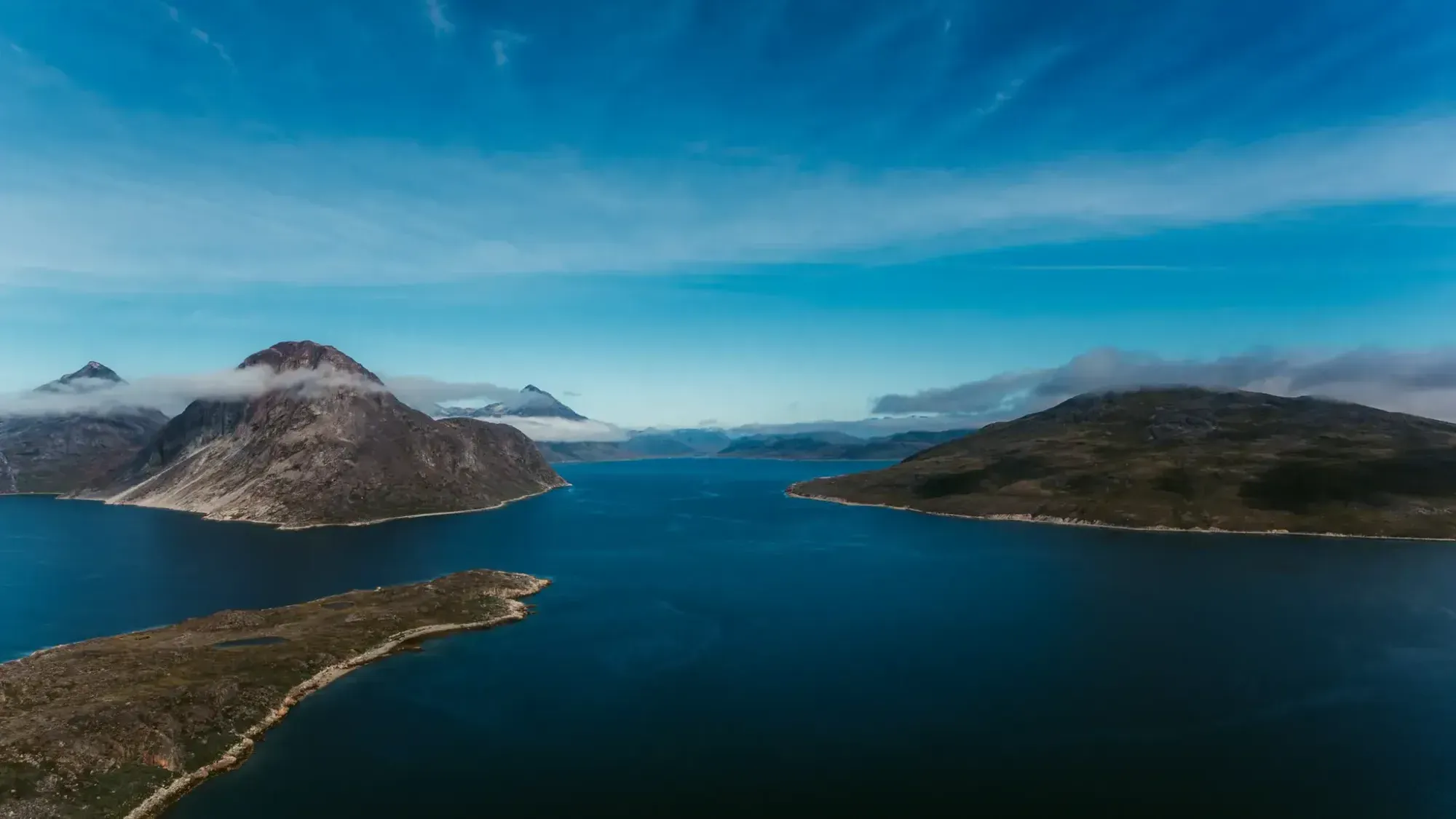 A wide fjord stretches between rocky, low-lying islands and mountains under a partly cloudy blue sky, captured in Greenland by Chris Zielecki