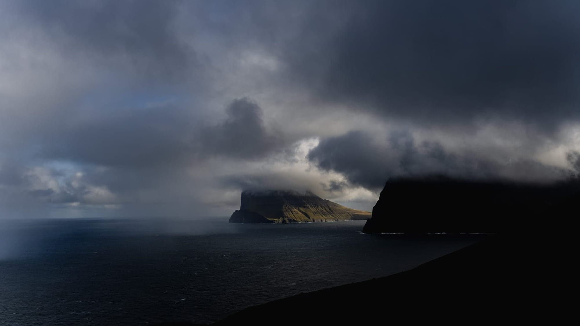 Dramatic storm clouds embrace rugged coastal cliffs in this moody seascape from the Faroe Islands, masterfully captured by elopement photographer Sturmsucht